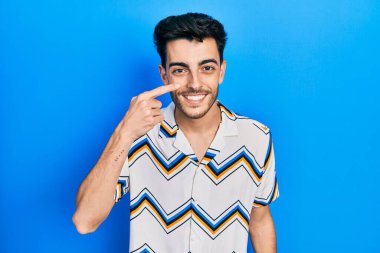 Latin asıllı genç bir adam, gündelik giysiler giyip el ve burnu işaret ediyor, neşeli bir şekilde gülümsüyor. güzellik konsepti 