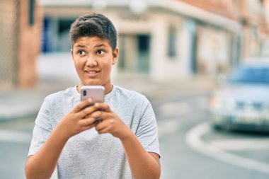 Sevimli Latin çocuk şehirde akıllı telefon kullanarak mutlu bir şekilde gülümsüyor..