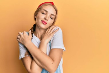 Güzel sarışın, beyaz kadın, günlük tulumlar giyip kendine sarılıyor, mutlu ve pozitif, kendinden emin gülümsüyor. Sevgi ve özgüven. 