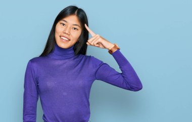 Güzel, genç Asyalı kadın günlük kıyafetler giyip gülümsüyor. Tek parmakla kafasını işaret ediyor. Harika bir fikir ya da düşünce, iyi bir hafıza. 