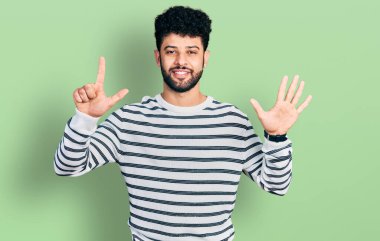 Sakallı, sıradan çizgili süveter giyen genç Arap adam yedi numaralı parmaklarını gösterip gülümserken kendinden emin ve mutlu.. 