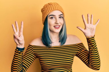 Genç modern kız yün şapka takıyor ve 8 numaralı parmaklarıyla işaret ederken kendinden emin ve mutlu gülümsüyor.. 