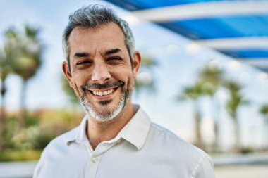 Orta yaşlı, kır saçlı, gülümseyen bir adam şehirde mutlu bir şekilde duruyor..