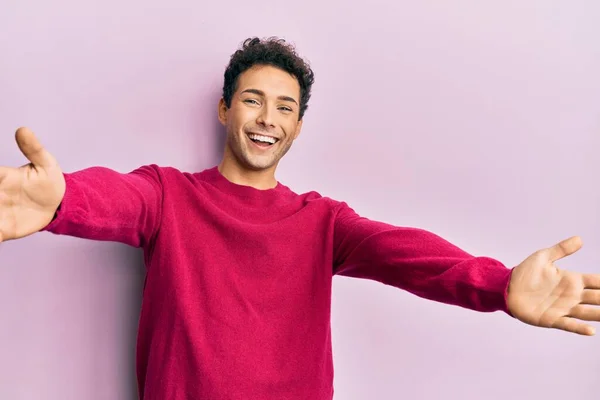 ピンクの背景にカジュアルな服を着たハンサムなヒスパニック系の男が抱擁のために腕を開いて笑顔カメラを見て 幸せを受け入れる陽気な表情 — ストック写真