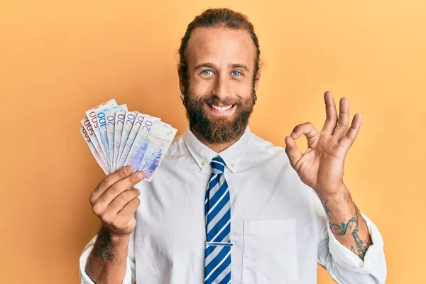 髭と長い髪を持つハンサムな男は 指でOkサインを行う甘いクローナ紙幣を保持し 笑顔優しいジェスチャー優れたシンボル — ストック写真