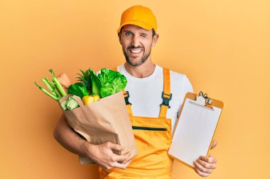 Genç yakışıklı, kurye üniforması giyen, market alışverişi yapan ve panoya göz kırpan seksi bir ifadeyle, neşeli ve mutlu bir yüzle bakan bir adam.. 