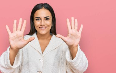 Genç esmer kadın gündelik giysiler giyer ve parmaklarını on numarayla gösterirken kendinden emin ve mutlu bir şekilde gülümser.. 