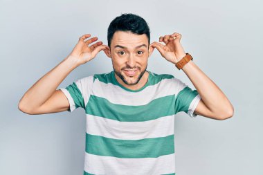 Günlük kıyafetler giyen, gülümseyen, parmaklarıyla kulaklarını çeken genç İspanyol bir adam, komik bir jest. deneme sorunu 