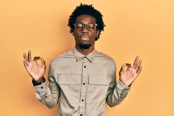 カジュアルな服と眼鏡を身に着けている若いアフリカ系アメリカ人の男性は 指で瞑想のジェスチャーを行う閉じて目でリラックスして笑顔 ヨガのコンセプト — ストック写真