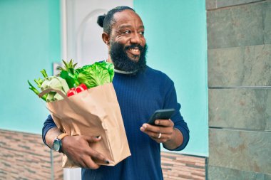 Sakallı Afrikalı Amerikalı adam elinde süpermarketten alınmış kağıt torbalarla akıllı telefon kullanıyor.