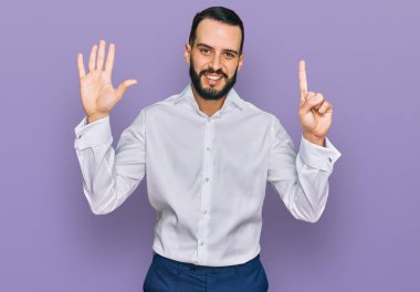 Sakallı genç adam iş gömleği giyiyor ve altı numaralı parmaklarıyla işaret ederken kendinden emin ve mutlu bir şekilde gülümsüyor.. 