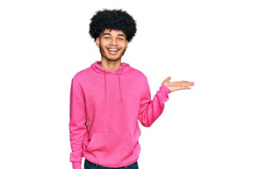 Afro saçlı, günlük pembe kazak giyen neşeli bir şekilde gülümseyen ve avuç içi kameraya bakan Afro-Amerikan genç adam.. 