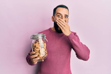 Sakallı genç adam elinde çikolata parçacıklı kurabiye kavanozuyla ağzını eliyle kapadı, şok oldu ve hata yapmaktan korktu. şaşırmış ifade 