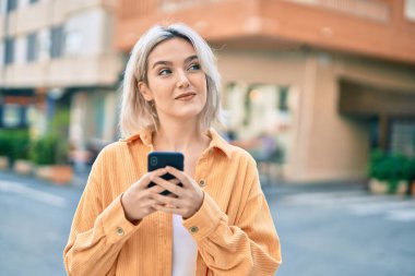 Genç sarışın kız şehirde akıllı telefon kullanarak mutlu bir şekilde gülümsüyor..