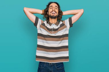 Genç İspanyol bir adam günlük kıyafetler giyiyor rahatlıyor ve esniyor, kolları ve elleri başının arkasında mutlu bir şekilde gülümsüyor. 