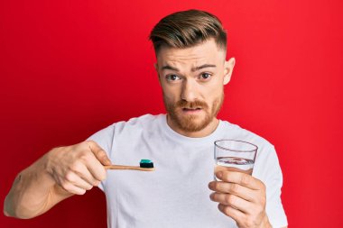 Kızıl saçlı genç adam diş fırçasıyla diş macunu ve bir bardak sudan habersiz ve kafası karışmış bir ifade tutuyordu. şüphe kavramı. 