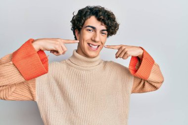 Günlük kışlık süveter giyen, neşeli gülümseyen ve parmaklarıyla ve ağzıyla işaret eden İspanyol genç adam. diş sağlığı kavramı. 