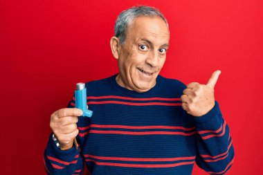 Tıbbi astım spreyini elinde tutan kıdemli İspanyol adam parmağını yukarı kaldırıyor ve ağzı açık gülümsüyor. 