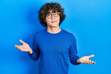 Sıradan kıyafetler giyen, gözlüklü, kafası karışmış, elleri ve kolları havada yakışıklı bir genç. şüphe kavramı. 