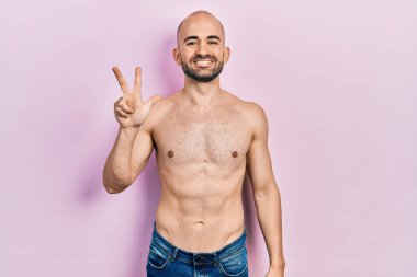 Genç kel adam üstsüz duruyor ve üç numaralı parmaklarıyla işaret ediyor kendinden emin ve mutlu bir şekilde gülümsüyor.. 