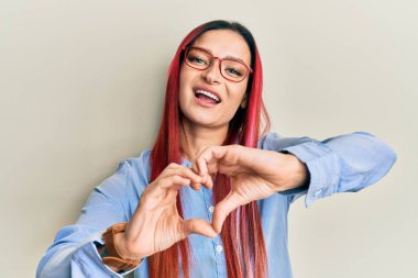 Gündelik giysiler ve gözlük takan beyaz bir kadın aşıkken gülümsüyor. Elleriyle kalp sembolü yapıyor. romantik kavram. 