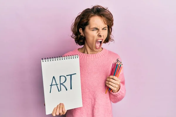 アートノートを持っている若いブルネットの女性と色の鉛筆怒っていると怒っている叫び不満と激怒し 怒りで叫んでいる 怒りと攻撃的な考え方 — ストック写真