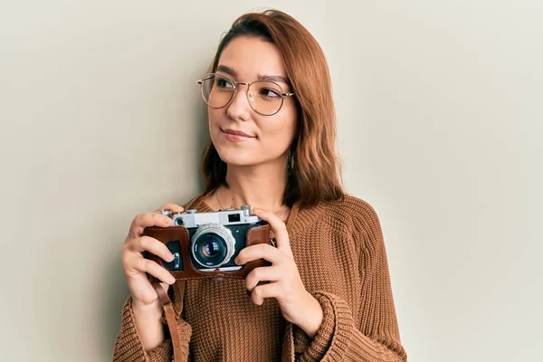 Young caucasian woman holding vintage camera smiling looking to the side and staring away thinking.