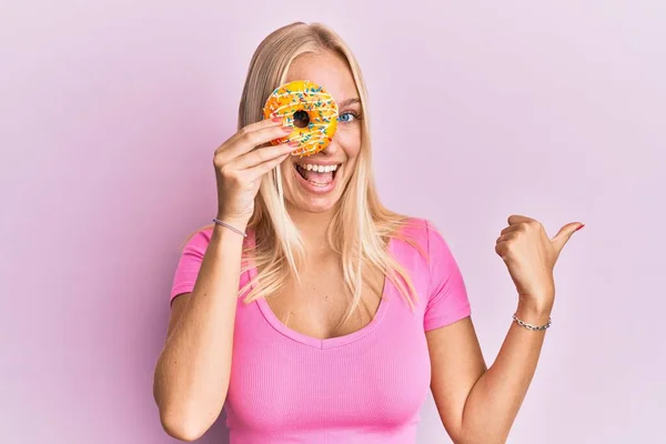 Young Blonde Girl Holding Donut Pointing Thumb Side Smiling Happy — ストック写真