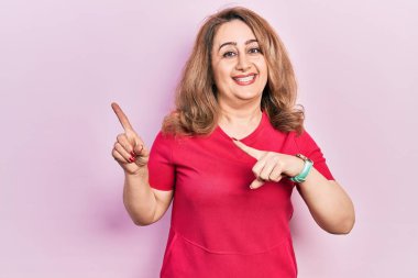 Orta yaşlı beyaz kadın günlük kıyafetler giyiyor gülümsüyor ve kameraya bakıyor iki eliyle ve parmaklarıyla kenarı işaret ediyor. 