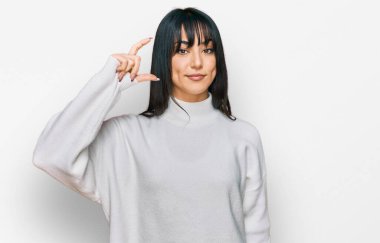 Kâküllü genç esmer kadın balıkçı yaka kazak giyiyor gülümsüyor ve kendine güvenen el hareketleriyle küçük beden işareti yapıyor parmakları bakıyor ve kameraya bakıyor. ölçüm kavramı. 