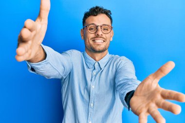 Gündelik giysiler ve gözlük takmış genç İspanyol adam kameraya bakıp sarılmak için kollarını açmış gülümsüyor. Mutluluğu kucaklayan neşeli bir ifade.. 