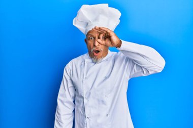 Orta yaşlı, gri saçlı, profesyonel aşçı üniforması giyen ve şapka takan, şaşırmış yüz ve gözlerle parmaklarından bakan bir adam. İnançsız ifade. 