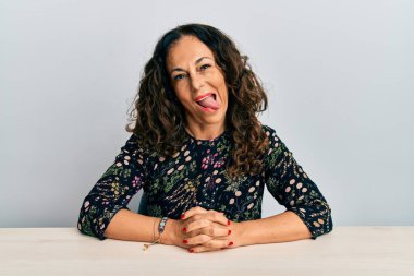 Orta yaşlı, güzel bir kadın, gündelik giysiler içinde masada oturmuş, komik bir ifadeyle dilini dışarı çıkartıyor. duygu kavramı. 