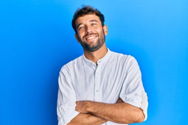 Sakallı, zarif tişörtlü, mutlu yüzlü, kollarını kavuşturup kameraya bakan yakışıklı bir adam. pozitif kişi. 