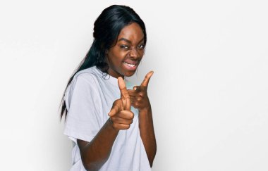 Gündelik beyaz tişört giyen genç Afro-Amerikalı kadın mutlu ve komik yüzlü parmaklarını kameraya doğrultuyor. iyi enerji ve titreşimler. 