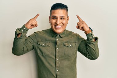 Gündelik kıyafetler giyen genç latin adam gülümseyerek iki eliyle kafasını işaret ediyor, harika fikir ya da düşünce, iyi hafıza 