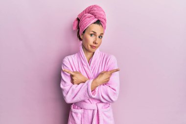 Duş başlığı ve bornoz takan genç sarışın kadın iki tarafı da parmaklarıyla işaret ediyor, farklı yönlere bakıyor. 