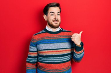 Sakallı, zarif yün süveter giyen, gülen yüzlü ve baş parmağıyla yüzünü gösteren yakışıklı beyaz bir adam.. 