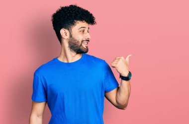 Sakallı, sıradan mavi tişörtlü, mutlu yüzlü ve baş parmağıyla yüzünü gösteren genç bir Arap.. 