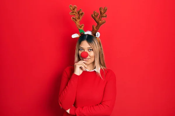 鹿のクリスマスの帽子と赤い鼻を身に着けている美しいヒスパニック系の女性は 交差腕と手で笑顔でカメラを見て自信を持ってあごに提起 ポジティブな — ストック写真