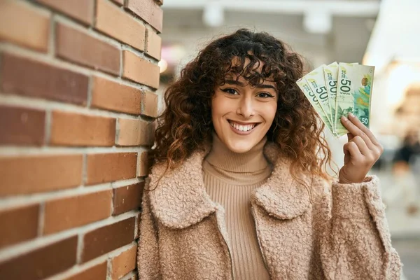 Young hispanic woman smiling happy holding israel shekels banknotes at the city