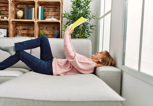中年金发女人开心地微笑着 躺在沙发上看书 — 图库照片