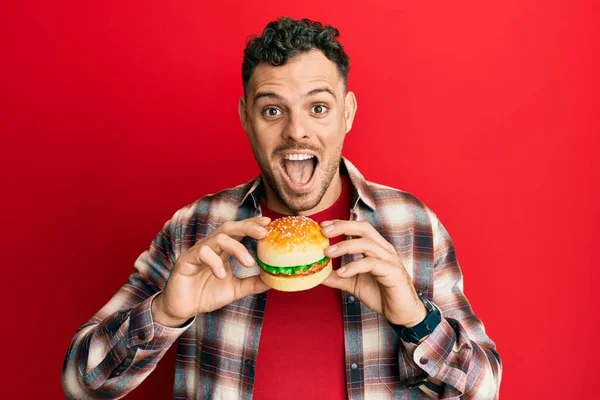 Młody Hiszpan Jedzący Smacznego Hamburgera Świętującego Szaleństwo Zdumiony Sukcesem Otwartymi — Zdjęcie stockowe