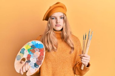 Güzel beyaz kız elinde fırça ve boya paleti tutarken yüzünde ciddi bir ifadeyle rahatladı. Basit ve doğal bir şekilde kameraya bakıyorsun.. 