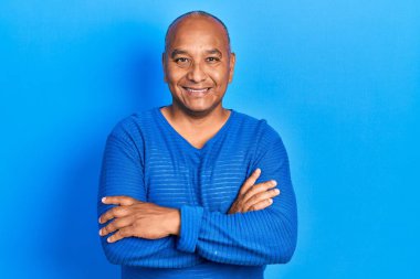 Orta yaşlı, sıradan kıyafetler giyen mutlu yüzlü, kollarını kavuşturup kameraya bakan Latin bir adam. pozitif kişi. 