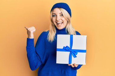 Genç beyaz bir kadın elinde hediyeyle baş parmağını kaldırmış, ağzı açık bir şekilde gülümsüyor. 