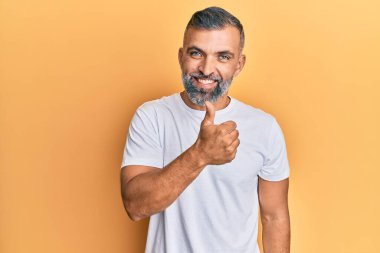 Orta yaşlı, beyaz tişört giyen yakışıklı bir adam mutlu bir el hareketi yapıyor. Kameraya bakıp başarıyı gösteren ifade. 