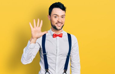 Hipster sakallı İspanyol adam papyon takıyor ve pantolon askısı takıyor. Beş numaralı parmaklarıyla işaret ediyor. Kendinden emin ve mutlu bir şekilde gülümsüyor.. 
