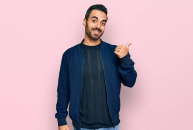 Gündelik kıyafetler giyen, gülümseyen, gülümseyen ve başparmağıyla yan tarafı gösteren İspanyol genç adam.. 