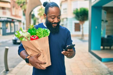 Sakallı Afrikalı Amerikalı adam elinde süpermarketten alınmış kağıt torbalarla akıllı telefon kullanıyor.